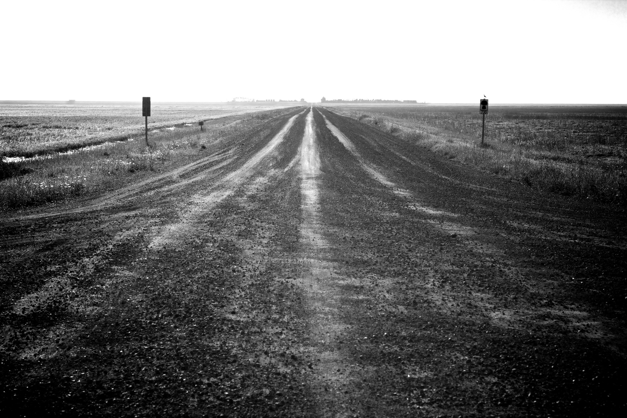 A black and white photo of a road crossing, with dirty roads in all directions. We se a city far off in the distance.