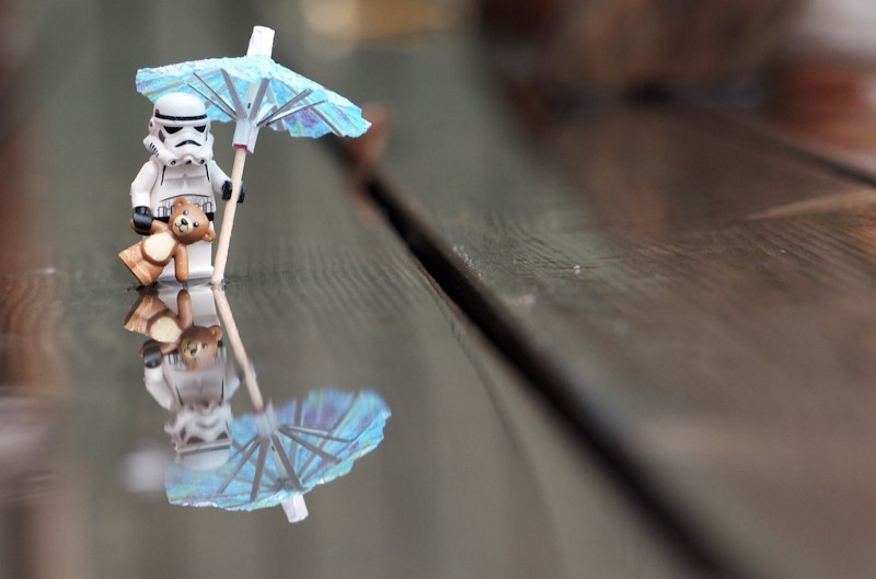 A Star Wars storm trooper lego figure carrying a brown teddy bear and a blue umbrella. They are on top of a wooden, wet, surface.