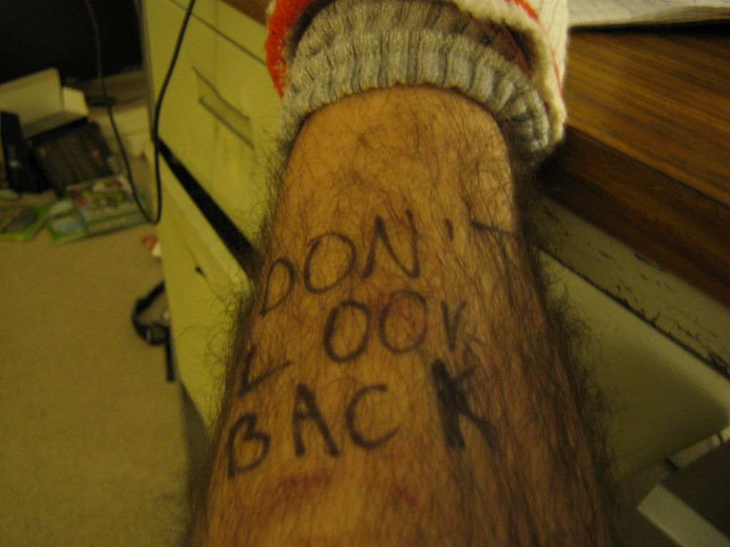 A picture from a dirty and messy room, we see a leg on top of a desk. The pants pulled up to show a hairy white persons leg with the words "Don't look back" written on it