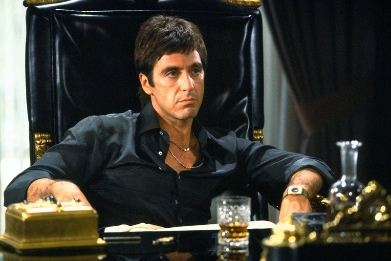 A picture of the character Tony Montana, from the movie Scarface. Sitting at an office desk. There is a drink and gold items on the