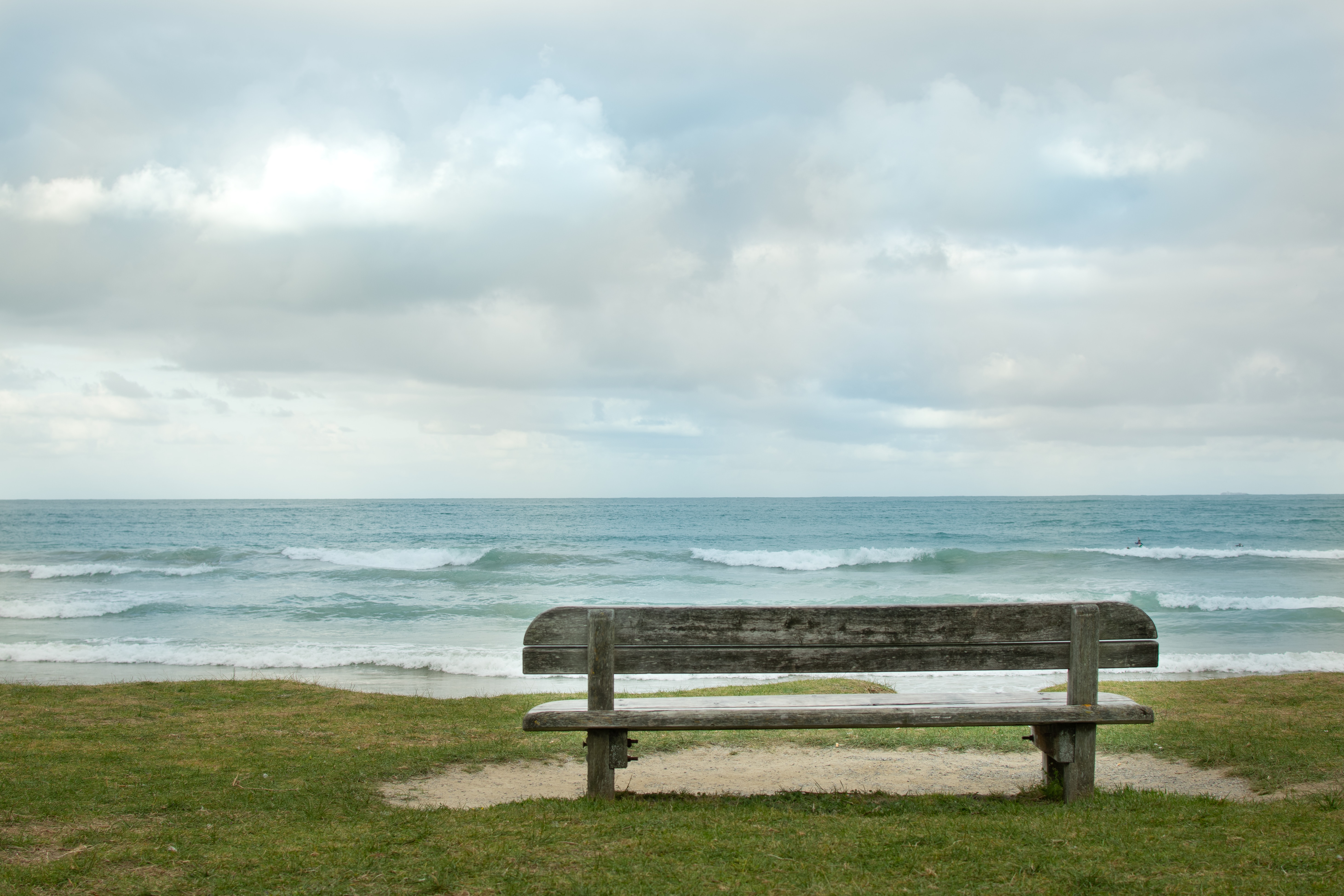 A bench on the grass in front of a white sandy beach. The ocaen is in the distance. There are waves coming in.