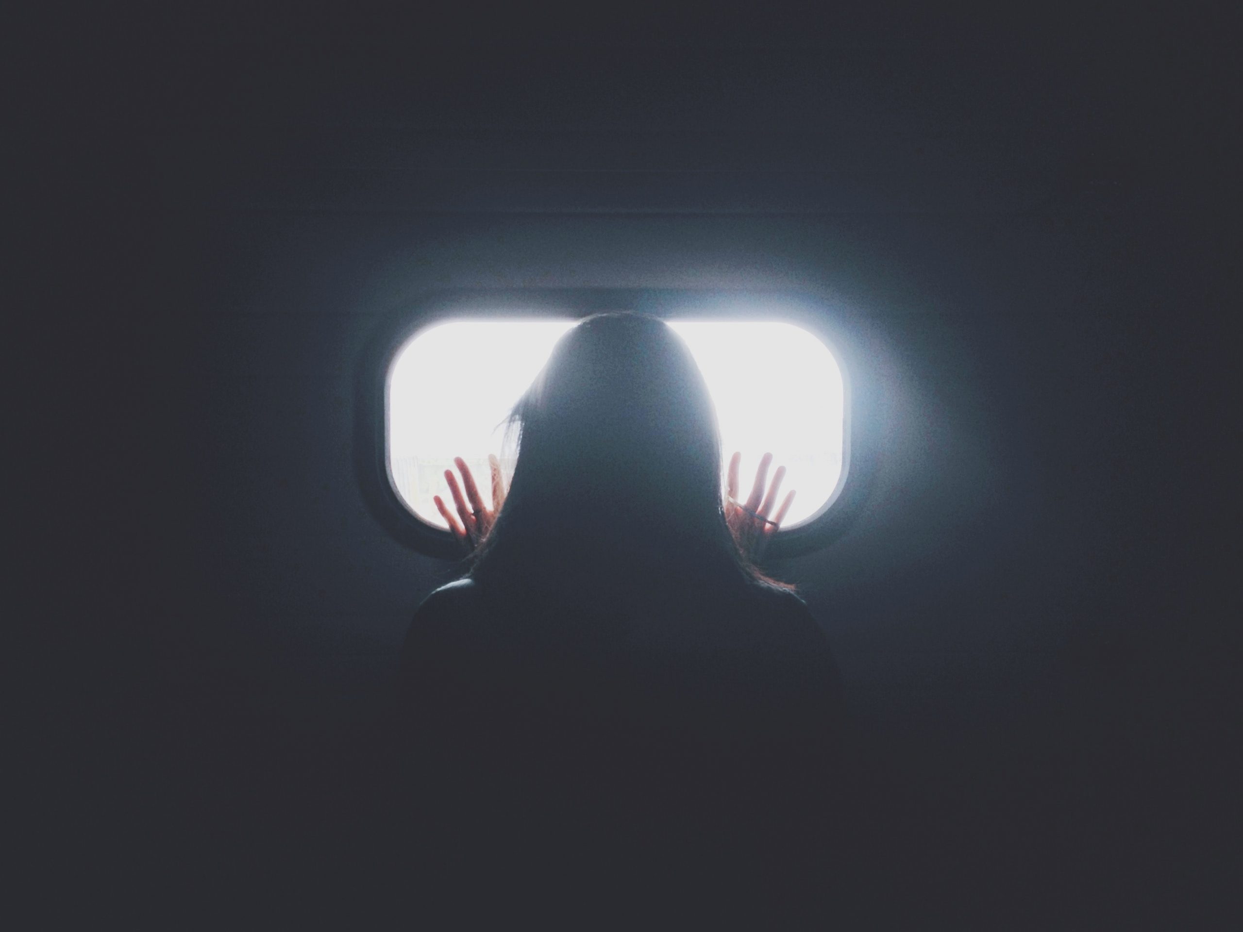 Person with long hair in a dark room looking through a window with light coming through it.