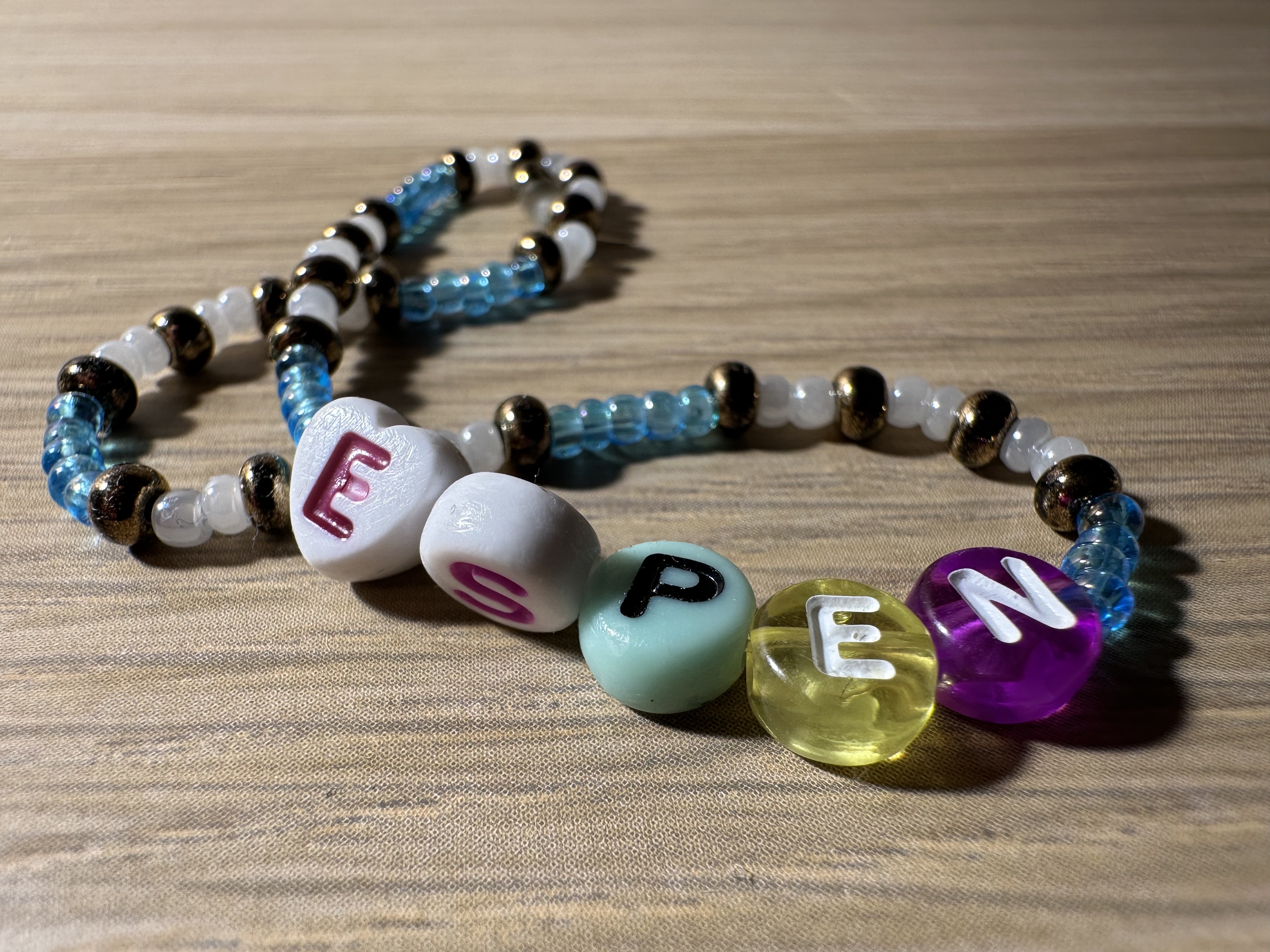 A colorful bracelet laying on a wooden surface. The letters e, s, p, e, n are attached to the string.