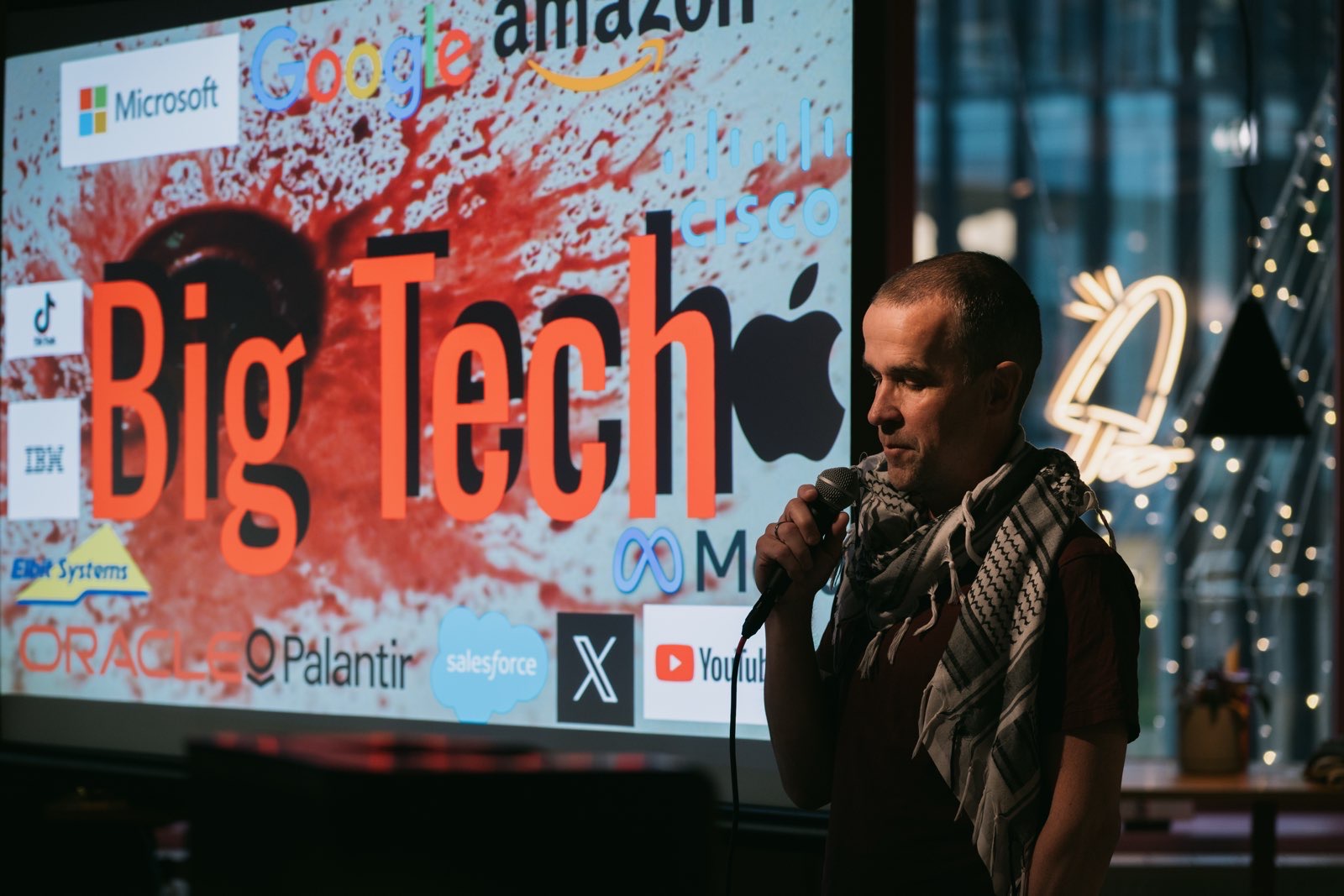 Me stadning with a microphone in my hand, wearing the keffiey, in front of a slide with the words Big Tech in red letters. In the background is a sink with blood and on top company logos from companies like Google, Meta, AWS, etc